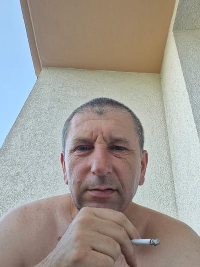 A close-up, low-angle, selfie-style shot shows a fair-skinned, middle-aged Caucasian male with short, dark hair with some graying. He is shirtless, with his chin resting on his right hand, and holding a lit cigarette in his left hand. His expression is neutral, and his eyes are looking directly at the camera. He appears to be outdoors, with a light blue sky and a beige overhang visible in the background, suggesting a balcony or an exterior architectural feature. The wall behind him has a textured, speckled finish, typical of exterior stucco. The lighting is bright, indicating daytime. There is no identifiable information to pinpoint this scene to a specific location in Brno, Czechia.
