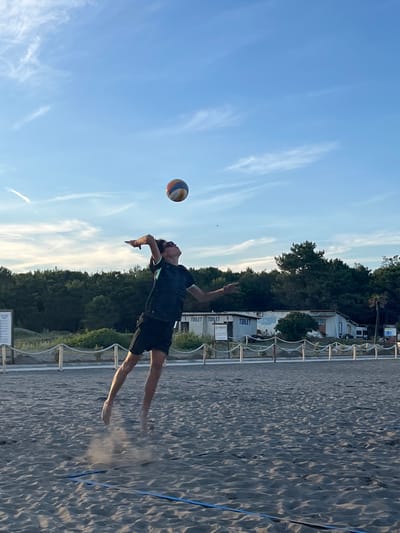 A young person is captured mid-air on a sandy beach court, executing a volleyball serve. The player, wearing a dark t-shirt with a lighter trim and black shorts, is focused on the ball suspended above and slightly in front of them. Their left arm is extended upwards, and their right arm is brought back, poised to strike. The sand is kicked up around their feet, indicating the power of their jump. In the background, a line of trees forms a dense green backdrop against a clear blue sky with wispy white clouds. Several small, white buildings with "TOILET" signs are visible behind a rope fence that separates the court from the background area. The beach volleyball court has visible blue boundary lines on the sand. The scene appears to be set in the city of Pistula, Montenegro, during daylight.