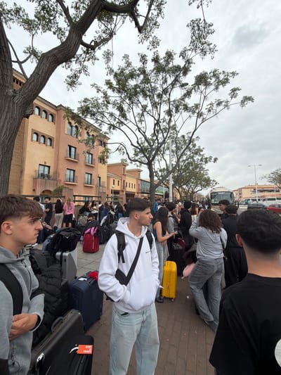 In Punta Umbría, Spain, a large group of young people with luggage is waiting outdoors. They are sitting or standing on a brick-paved area, near a building with multiple stories and a light orange facade. A large tree with thin branches and sparse leaves stands to the left of the frame. In the background, a bus can be seen, suggesting the people are waiting for transportation. The sky is overcast.