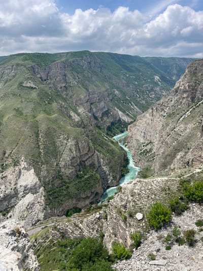 The image depicts a steep canyon landscape with a turquoise river running through it. The canyon walls are a mix of rock formations and green vegetation. In the distance, there are mountains covered in lush greenery under a sky with scattered clouds. Along the cliff edge, a road can be seen. There are no people visible in the image. The location may be near the city of Druzhba, Russia, given the user's request.