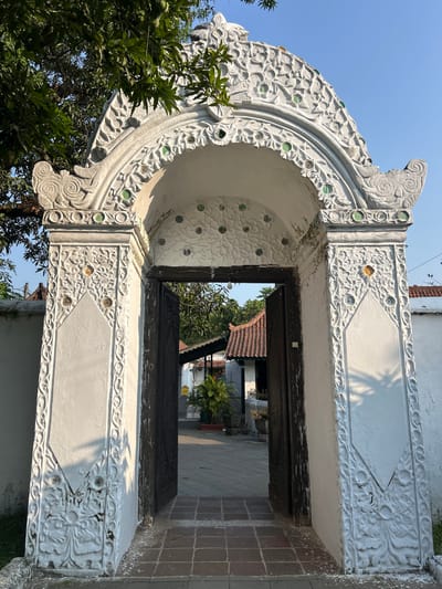 The image depicts the intricately carved white archway of the Sunan Gunung Jati tomb complex in Cirebon, Indonesia. The archway is adorned with floral motifs and embedded with small green glass circles, while the door appears to be made of dark wood and is partially open. Beyond the archway, a courtyard is visible with traditional Indonesian tiled roofs and lush greenery. The sun casts shadows on the white walls of the complex, indicating it is daytime.