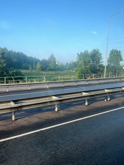 The image shows a view of a highway with green trees and a light blue sky in the background. The highway has metal guardrails separating lanes. The road surface is dark asphalt with a white line marking a lane. In the distance, there is a line of trees, some of which appear to be deciduous, and a faint mist hangs in the air. There is a street lamp on the right side of the image. The perspective suggests the photo was taken from a vehicle moving along the highway. The scene is likely in a suburban or rural area of Maardu, Estonia, given the highway infrastructure and the presence of abundant greenery. No people are visible in the image.