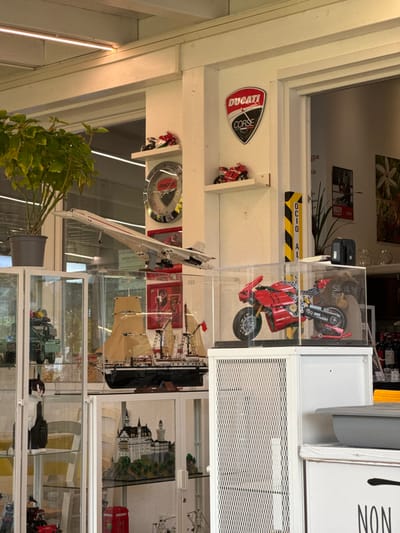 The image displays an indoor area in Anzola dell'Emilia, Italy, characterized by white walls and a white wooden beamed ceiling equipped with strip lighting. No individuals are visible.
In the upper central portion, a large white model of a Concorde aircraft is prominently displayed, positioned in flight below a potted green plant. On the adjacent white wall, a circular metallic emblem bearing the "DUCATI" logo is mounted. Above this emblem, a small red and white model motorcycle rests on a white shelf, with two similar miniature motorcycles also present on shelves to the left. A "DUCATI CORSE" shield logo is affixed to the wall further right, positioned above another small red model motorcycle on a shelf. A yellow and black vertical post with partial text "OCIO AL" is visible through a doorway on the right.
The lower section of the image features multiple white-framed glass display cases. A central case contains a detailed model of a three-masted sailing ship with a black hull, tan sails, and a red flag. Below this, another case exhibits a detailed model of a white castle on a green base, alongside a small red model of a British telephone booth. A partially visible case on the far left contains a green model vehicle and a black and white cat figurine. In the foreground right, a clear acrylic display case houses a detailed red model of a Ducati motorcycle on a stand. This display rests on a white cabinet with a perforated metal front. To its right, another white cabinet is partially visible, topped by a gray rectangular container, and displays the partial text "NON" on its front.