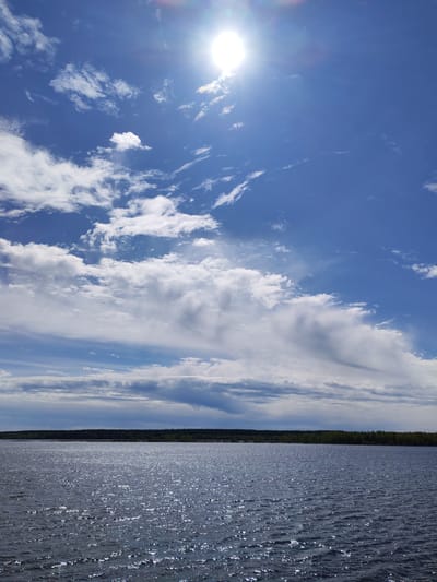 The image shows a body of water, likely a lake or sea, with the opposite shore visible in the distance. The shoreline is densely lined with trees, creating a dark green strip against the horizon. The water is choppy with visible ripples and highlights, reflecting the sunlight. Above the water, the sky is a vibrant blue, filled with white clouds of varying shapes and sizes. The sun is positioned near the top of the frame, appearing as a bright, circular light source. The location may be in Baburino, Russia.