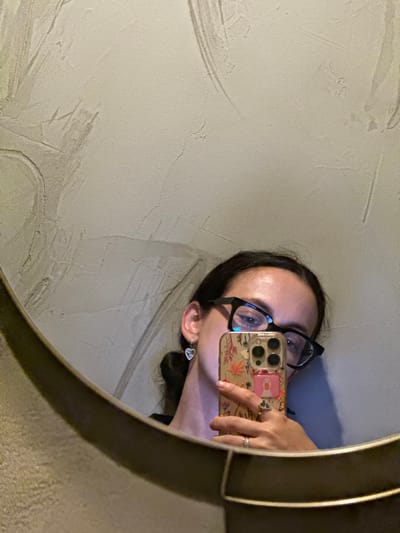 A young woman with dark hair pulled back is reflected in a large, oval mirror. She is wearing black-rimmed glasses and a heart-shaped earring. Her right hand is holding a smartphone with a floral case and a pink duck-shaped grip. She is also wearing two rings on her right ring finger. The background of the reflection is a textured, light-colored wall. The mirror is framed by a dark, metallic rim. The lighting is dim, suggesting an indoor environment.