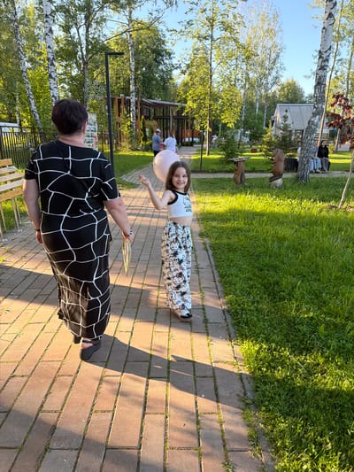 The image depicts an outdoor scene in what appears to be a park or recreational area in Zaprudnya, Russia, bathed in warm, low-angle sunlight, indicating either morning or late afternoon.

In the foreground, a woman is walking away from the camera on a light-brown brick-paved path. She has short, dark hair and is wearing a black dress or long top with a white abstract geometric pattern. She holds a thin, gold-colored rectangular object in her right hand and wears black open-toed platform sandals. A black strap is visible over her left shoulder. Behind her, slightly to the right, a young girl, approximately 6-8 years old, walks towards the camera on the same path, smiling. She has long, dark brown hair and is holding a light pink, spherical balloon by its string in her left hand. The girl is wearing a white sleeveless crop top with black trim and a "Hello Kitty" logo, along with white trousers patterned with "Hello Kitty" characters and text. She wears black and white shoes.

To the right of the paved path, there is a manicured grassy area. Two animal-like sculptures, resembling beavers or groundhogs, are visible on the grass. One is holding a flat, rectangular object on its head. Further back on the right, two individuals are seated on a bench under a tree.

The background is densely treed, primarily with birches identifiable by their white bark, and other deciduous trees with green foliage. Several wooden structures, including a small house-like building with a light-colored roof, are scattered throughout the park. On the left side of the path, behind the woman, a black lamppost or decorative light fixture featuring a white snowflake-like ornament is visible, with a metal fence behind it. Several other indistinct figures can be seen walking or congregating in the deeper background. Long shadows are cast across the paved path and grass, originating from the left side of the frame.
