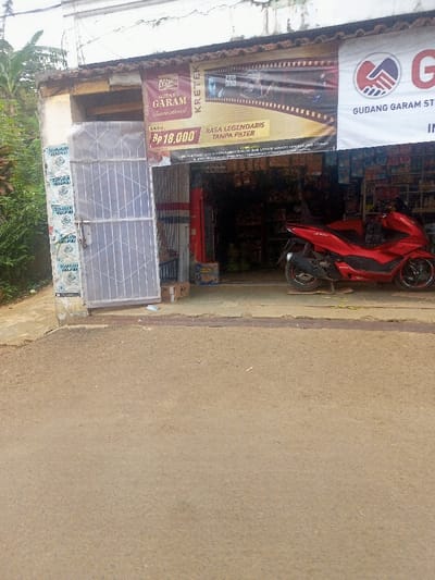 This street-level view captures a small, humble shop in Cimaung, Indonesia, with a weathered facade. In the foreground, a dusty, light-brown road or path occupies the lower half of the image, leading up to the shop's entrance.

The shop's entrance features a metal rolling gate, partially open, revealing a dark interior. A separate, white metal gate with a crisscross pattern is also partially ajar on the left side, secured by a padlock. The wall pillar to the left of this gate is covered with numerous old, crumpled white papers, possibly remnants of flyers or notices.

Above the entrance, large advertising banners are prominently displayed. The most visible banner promotes "GUDANG GARAM International" cigarettes, identified as "KRETEK." It shows a price of "Rp 18,000" and the slogan "RASA LEGENDARIS TANPA FILTER" (Legendary Taste Without Filter). Another banner, partially visible on the right, also bears the "GUDANG GARAM ST" logo and name.

A bright red scooter, resembling a Honda PCX, is parked in front of the shop, facing right. A few cardboard boxes are visible near the shop's threshold. To the far left, lush green vegetation, possibly bushes or trees, can be seen beyond the building. The scene appears quiet and devoid of people, suggesting a tranquil moment during daytime, possibly under overcast skies given the diffused lighting and lack of harsh shadows.
