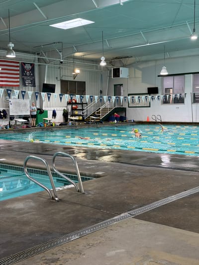 The image depicts the interior of an indoor swimming pool facility, likely in Johns Creek, Georgia, United States, given the presence of US flags and the general characteristics of such facilities. The pool is rectangular and has multiple lanes, delineated by lane lines. Swimmers are visible in the pool, engaged in swimming activity. Poolside, individuals appear to be supervising or spectating. The facility includes features such as a staircase leading to an elevated platform, signage, and equipment storage. Overhead, the structure includes lighting fixtures and structural support beams. The surrounding walls are primarily white and there are windows and televisions.