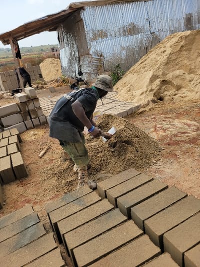 The image depicts an outdoor construction or brick-making site in Nwofia, Nigeria, under an overcast sky. In the foreground, a male individual, observed from a three-quarter back view, wears a dark t-shirt, a dark apron-like garment, camouflage-patterned trousers, and a faded green/brown bucket hat. He is actively engaged in scooping loose, light brown granular material from a pile on the reddish-brown earth using a small, light-colored spade into a yellow container held in his left hand. A blue bracelet is visible on his right wrist.
Numerous light brown, rectangular bricks are neatly stacked in organized rows in the immediate foreground and extending to the left side of the frame. In the left background, a second male figure, partially obscured, is visible stacking similar bricks near a corrugated metal shelter with a wooden-framed roof overhang. Adjacent to this shelter and behind the central worker is a significant mound of light brown sand. The ground consists of bare reddish-brown earth mixed with scattered granular material and small wood fragments. In the distant background, low-lying vegetation and the upper portion of a partially constructed building are discernible.