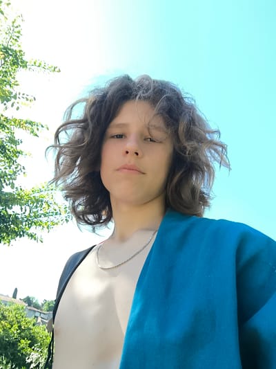 A person with curly brown hair and a silver necklace is looking at the camera from a low angle. The person's chest is exposed, with a black strap on the left and a blue fabric draped over the right shoulder. Behind the person is a bright blue sky. To the left of the person, there is a tree with green leaves. In the background, there are rooftops of buildings and green foliage, suggesting a location in Chézenas, France.