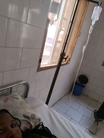 In a hospital room located in Mape, Nigeria, an individual with dark skin and braided hair is partially visible in the bottom-left, lying on a hospital bed equipped with a metal headboard, white sheets, and a floral-patterned pillow. The person's face is turned towards the camera. An intravenous (IV) pole stands prominently in the mid-ground, supporting two clear bags of IV fluid. One IV line descends from the rightmost bag towards the patient, indicating ongoing medical treatment. The room features white square tiled walls and a light-colored tiled floor. A window with a light-colored curtain and a visible wooden frame allows bright natural light into the room. In the bottom-right corner, a blue bucket with a dark lid, likely a waste receptacle, is situated next to a foot clad in a light-colored sneaker.