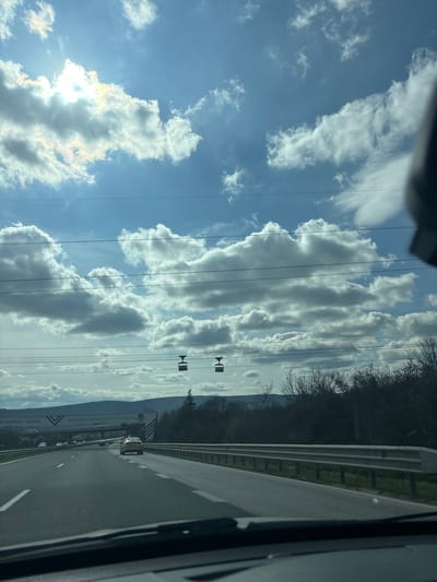 The image shows a view from a vehicle traveling on a highway in Kipra, Bulgaria. To the right of the road is a metal guardrail and a tree-lined area. Overhead power lines traverse the sky, which is filled with puffy white clouds against a blue background. In the distance, a cable car system is visible. The road has multiple lanes and white lines. A billboard with the name “ВАРНА” is visible on the side of the road. There are other cars traveling on the road. The view is slightly obscured by the vehicle frame on the right edge of the picture.