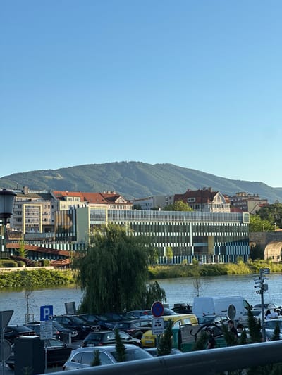 The image captures a detailed urban and natural landscape in Maribor, Slovenia, under a clear, bright blue sky. The scene is observed from an elevated perspective overlooking the Drava River and its surroundings.
In the immediate foreground, a dark metal railing runs horizontally across the bottom of the frame, suggesting the viewpoint is from a bridge or elevated platform. Below the railing is a paved parking lot densely occupied by various vehicles, including sedans, SUVs, and vans of different colors (e.g., yellow, white, black). Visible signage includes a blue "P" parking sign indicating "CONA 4" (Zone 4) with operational hours and Slovenian text "plačljiv park. na območju parkirišča" (paid parking in the parking area). Another red circular prohibition sign with a diagonal line and a pictogram of a truck/van is also present. Several individuals are visible within the parking lot, some standing near vehicles, others walking. On the right, a pole equipped with multiple security cameras monitors the area. Lush green vegetation, including a large weeping willow tree, partially borders the parking lot and riverbank.
The midground features the calm, blue-grey surface of the Drava River. On the opposite bank, green foliage lines the water's edge. A prominent, large, modern multi-story building dominates the central midground. Its facade is characterized by a pattern of vertical alternating dark and light (light blue/white) striped panels and large reflective windows. This building is identifiable as part of the University of Maribor, likely the Faculty of Electrical Engineering and Computer Science (FERI). Behind and to either side of this modern structure, several older, multi-story buildings with diverse architectural styles, including red-tiled pitched roofs and varying facade colors (e.g., yellow, grey), are visible. One particularly ornate, traditional European-style building with a prominent roof and multiple windows is discernible on the right side behind the modern complex. Further to the right, a stone arch structure, potentially part of a historic bridge or embankment, is partially visible.
The background is dominated by a large, heavily forested mountain range, identified as Pohorje. The mountainside exhibits patches of cleared land or trails, possibly indicative of ski slopes or logging. A communication tower or antenna structure is clearly visible on one of the higher peaks of the range. The sky above is entirely clear and light blue, devoid of clouds.