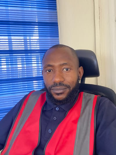 This is a close-up, rotated portrait of a Black man seated in a chair in front of a blue vertical blind. He has a beard and is wearing a dark blue collared shirt with a red and gray high-visibility vest over it. The man's face is partially visible, with his eye and mouth in focus. The lighting suggests it is daytime, and the environment appears to be indoors, possibly an office or a workspace, given the presence of office furniture and blinds. There is no visible text in the image. The man's expression is neutral.