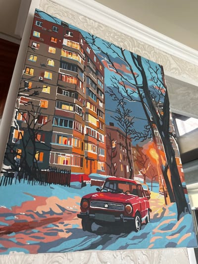 A photograph captures a canvas painting depicting a winter urban scene in Chernomurovskii, Russia, at dusk or night. The painting, rendered with distinct color blocks typical of a paint-by-numbers style, shows a red vintage-style sedan, possibly a Lada, parked on a snow-covered street in the foreground. Patches of snow with warm orange reflections from a streetlamp cover the ground. In the midground, bare trees with dark branches stand against a blue-grey sky with faint pink and orange clouds. A bright yellow-orange streetlamp illuminates the scene to the right. In the background, a multi-story residential building, characterized by numerous illuminated windows displaying warm orange and yellow lights, dominates the left side. Another, less prominent building is visible further back. The overall environment suggests a cold, settled evening in a Russian city. The painting rests against a white wall with decorative crown molding visible at the top of the photographic frame.