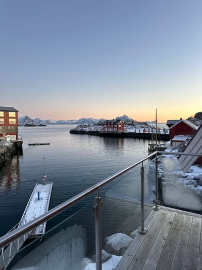 The image presents a panoramic view of a winter coastal scene in Stamsund, Norway, observed from a wooden deck featuring a glass and stainless steel railing in the foreground. The railing’s stainless steel posts and top rail are prominent, with large icicles and snow visible on the rocks immediately below and beside the deck. A calm, dark body of water occupies the midground. To the left, a four-story red building with multiple illuminated square windows stands on the water's edge, connected by a small floating gangway. In the center-left, a snow-covered white floating dock with a blue cylindrical post and a red lifebuoy on a vertical pole is present in the water. Across the water, on a snow-dusted landmass, a distinct red house with white window frames and a chimney is positioned. To its right, a sailboat with a tall mast is moored. Further right, partially obscured by the foreground, another red building with a sloped, snow-covered roof and an attached wooden structure is visible. The background consists of a chain of snow-capped mountains under a clear sky that transitions from a light blue overhead to a warm orange and pink hue at the horizon, indicative of either sunrise or sunset. No people or animals are depicted.