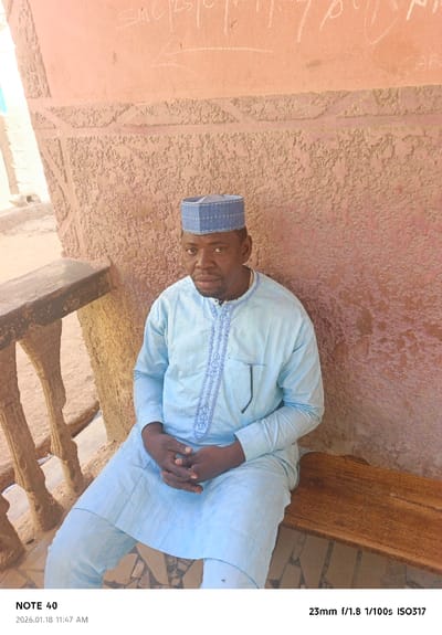 An adult male is depicted seated on a wooden bench in Tudun Wazurchi, Nigeria. The man, with dark skin, wears a light blue long-sleeved kaftan featuring light blue embroidery along the chest and collar, and matching light blue trousers. He has a dark-framed object, possibly eyeglasses, tucked into his left breast pocket. On his head is a blue cap with a light-colored geometric pattern. His hands are clasped in his lap. Behind him is a textured wall, colored in hues of light reddish-brown, exhibiting horizontal decorative bands and rough plasterwork. White chalk-like markings, including "SMC" and an arrow pointing left, are visible on the upper portion of this wall. To the man's left, a light-colored balustrade or railing with intricate, possibly carved, vertical supports is present. The ground beneath the bench and to the left of the man displays an irregular pattern of light and dark tiling or mosaic work. Natural daylight illuminates the scene.
