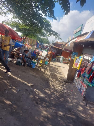 This image depicts a vibrant street market scene in Picha Ya Ndege, Tanzania. The foreground is dominated by a dusty, sun-drenched path, with dappled sunlight filtering through the leaves of trees overhead, suggesting it is daytime with clear skies.

Several people are present, engaged in various activities. In the center, a woman sits on a blue plastic chair, wearing a patterned dress and a headscarf, appearing to be attending to her wares or taking a break. Another woman in a floral dress is seated nearby, looking down at something in her hands. In the lower left, a man in a yellow t-shirt is visible from the chest down, his legs crossed, looking towards the market. Other individuals are sparsely scattered throughout the scene, some standing and others seated, suggesting a bustling but not overcrowded market.

The market stalls are laden with various goods. Colorful fabrics, possibly clothing or textiles, are displayed prominently on a stall in the upper right. Other stalls showcase what appear to be household items, food products, and possibly crafts. Blue and red tarps are visible, likely used for shade or to cover goods. There are also several plastic containers and buckets, hinting at produce or water being sold.

Visible text includes "Piga 150*60*#" on a red umbrella and some unreadable text on a blue and white sign in the background. The overall atmosphere is one of everyday commerce and community life in a tropical environment. The scene is lively and suggests a typical day at a local marketplace.