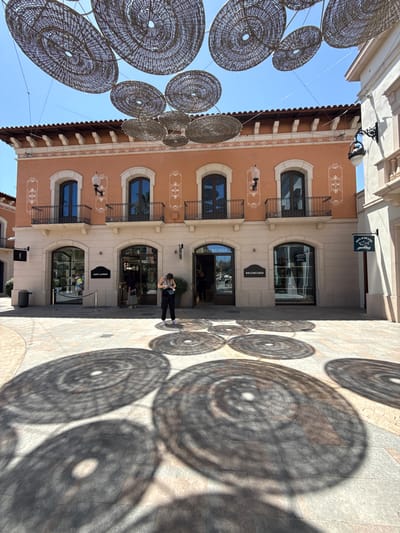 In La Roca del Vallès, Spain, a sunny day reveals an outdoor commercial area with a distinctive architectural style. A prominent orange building with arched windows and balconies stands as a backdrop, adorned with decorative white detailing. Above, large, circular woven structures are suspended, casting intricate circular shadows on the cobblestone ground.

The ground level features several retail establishments, including "Saint Laurent" and "Balenciaga," with glass storefronts offering glimpses of merchandise. A woman wearing a grey tank top and black pants is standing near the "Balenciaga" entrance, looking down at her phone. She is holding a small blue bag in her left hand. Other individuals are visible inside the stores, suggesting active commerce. The overall environment appears to be a pedestrian-friendly shopping district bathed in bright sunlight.