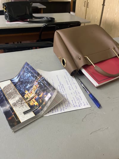 The image depicts a close-up of a desk with various items, likely in a classroom or study environment. In the foreground, a notebook with a scenic cover, featuring a mountain landscape and illuminated buildings, is open, revealing lined pages. Next to it lies a blue and black pen, positioned on a piece of paper filled with handwritten notes in Cyrillic script. A portion of a brown leather handbag is visible, with a glimpse of a red-covered book peeking out from within it. In the background, the edge of another desk and a black chair are discernible. The overall setting appears to be an indoor space with a light-colored desk surface and a wall with wooden cabinets behind it. There are no people visible in the image. The scene does not provide any specific indicators to pinpoint its location within Osh City, Kyrgyzstan.