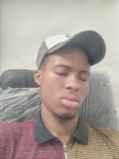 A single male subject is depicted in a medium close-up, seated indoors. The individual is wearing a grey and white baseball cap and a collared, horizontally striped shirt, maroon on the left side and yellow on the right, with a zipper closure. His eyes are closed, and his lips are slightly parted, exhibiting a glossy appearance. Minimal facial hair is visible around the chin. He appears to be resting or asleep. The subject is seated in a dark-colored chair, which is partially covered with clear plastic sheeting. The background is a plain, light-colored wall. The location of this scene is Kanti, Nigeria.