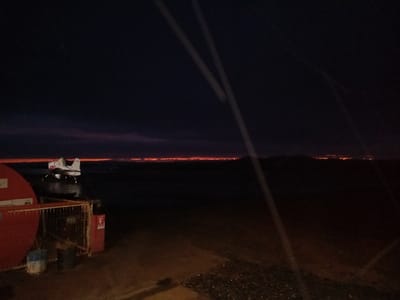 The scene, located in Santa Juana, Chile, captures a low-light environment at night or dusk. In the immediate left foreground, a large red cylindrical object, possibly a hose reel, is partially visible, alongside a metal grate enclosure containing indistinct objects and a small red sign. Centered in the midground, a white aircraft, identified by red markings on its tail and wing, rests on a dark, reflective surface, likely water, suggestive of an amphibious or floatplane design. The aircraft is oriented towards the right. The distant horizon presents a continuous, narrow band of intense orange-red light, extending across the frame, indicating significant distant illumination or a fire line. Above this, the sky darkens from a reddish-purple near the horizon to deep black overhead. Further right along the horizon, isolated clusters of orange lights are visible. Two faint, diagonal light streaks, consistent with lens flare or reflections, cross the upper image. No people are visible.