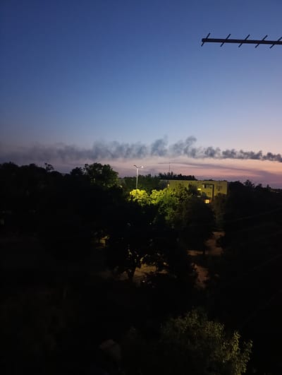 This is an outdoor, high-angle, full shot of a suburban or semi-rural scene at dusk. The sky transitions from a deep blue at the top to lighter hues of purple and pink near the horizon, indicating the end of the day. A dark, elongated antenna is visible in the upper right corner of the frame.
In the middle ground, a multi-story building with lit windows is nestled amongst dense, dark green foliage. A streetlamp illuminates a portion of the trees with a bright yellow light. Above the trees and the building, a long, dark plume of smoke drifts across the sky, creating a dramatic contrast against the fading light. The overall atmosphere is somewhat somber and quiet, possibly suggesting a peaceful evening or the aftermath of an event.
The presence of the building, trees, and streetlamp suggests a residential area. The smoke could be from a fire, industrial activity, or even a controlled burn, but its exact source is not discernable. The scene appears to be located in Ladyzhyn, Ukraine. There is no visible text in the image.