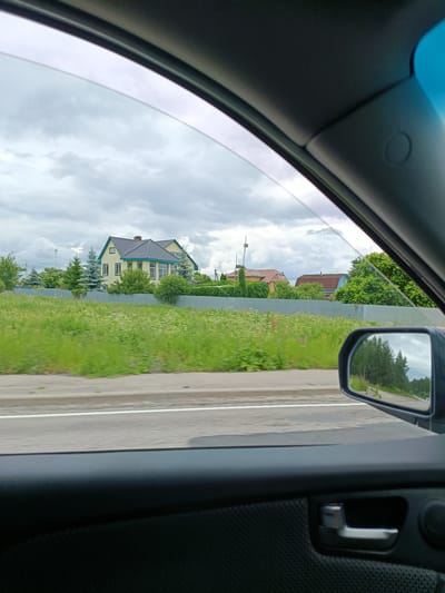The image captures a view from inside a vehicle, looking out through the side window and a side mirror. The foreground shows the interior of the car door with a dark textured panel and a silver door handle. Through the window, a rural landscape is visible under an overcast sky. There is a road with a white line marking in the middle distance, bordered by a grassy area with wildflowers. Beyond the grass, a grey metal fence runs horizontally, separating it from properties with houses and trees. One prominent yellow house with a dark roof and teal trim is visible, along with other buildings with red and blue roofs. The side mirror reflects a tree-lined road. The location is identified as Gribchikha, Russia. No people are visible in the frame.