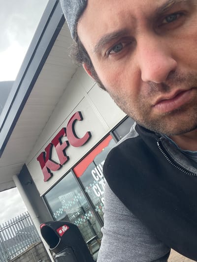 The image depicts a man taking a selfie in front of a KFC restaurant. The man is wearing a grey beanie, a grey hoodie, and a black vest. He is looking at the camera with a neutral expression. The KFC restaurant has its iconic red and white logo visible. A black trash can with red and white stickers is located to the left of the entrance. The background shows a grey, cloudy sky and some fencing. The overall scene suggests a casual, outdoor setting in a commercial area. The location is identified as Armagh, United Kingdom.