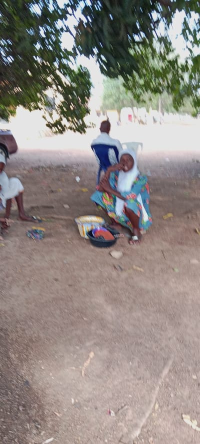 The image captures an outdoor scene, likely a communal or market area, under the shade of a large tree. The overall quality of the photograph is significantly blurry, suggesting camera shake or motion blur.
In the foreground, the ground is covered in light brown dirt or sand, with some scattered debris like small leaves and possibly bits of trash. A large, leafy tree canopy dominates the top of the frame, providing dappled shade and indicating a bright daytime setting.
There are at least three individuals visible. The most prominent figure is a woman positioned slightly to the right of the center, squatting or sitting very low to the ground. She is wearing a vibrantly patterned dress or tunic, predominantly blue with colorful floral or abstract designs in red, green, and white. Her head is covered with a light-colored, possibly white or cream-colored, headscarf or hijab. She is looking directly towards the camera with a wide, open-mouthed smile or laugh, conveying a cheerful and friendly demeanor. Her right hand is raised near her face. She appears to be barefoot, with a light-colored anklet visible on her right ankle. In front of her, on the ground, are two containers: a light-colored, possibly woven basket or rectangular container, and a dark, round bowl filled with various colorful items that are indistinct due to the blur but could be goods for sale or personal belongings.
Behind and to the right of this woman, a man is seated on a blue plastic chair, facing away from the camera. He is wearing a light-colored, possibly white or light blue, shirt over a darker garment. His posture is relaxed.
To the far left of the frame, only a partial view of another person is visible. This individual appears to be sitting low to the ground, possibly wearing a white garment, and their bare feet are visible. A small, dark object, possibly part of a vehicle or a bag, is also partially visible on the far left.
The background beyond the immediate gathering is very blurry but suggests an open area with possibly other distant trees, structures, or people. The activity appears to be an informal outdoor gathering, possibly a street vendor setting given the items in front of the central woman, characterized by relaxed interaction under the shade of a tree. No discernible text is visible in the image.
