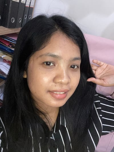 A person with dark hair is oriented at an angle, with their face close to the camera. The person has brown eyes, a fair complexion, and is smiling slightly. Their right hand is raised near their hair. They are wearing a black and white striped shirt. The background includes a pink surface and what appears to be a stack of black binders or files. The location is Negara, Indonesia.