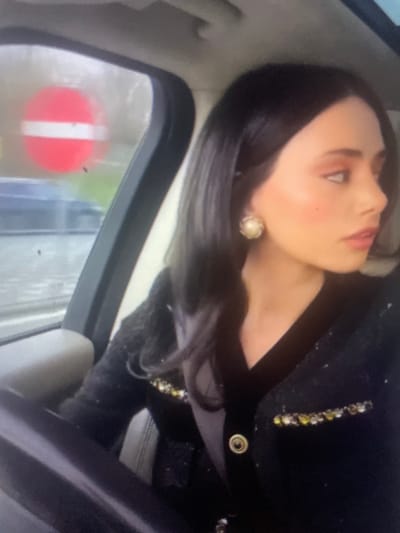 A young woman with dark hair and makeup is sitting in the driver's seat of a car. She is wearing a black jacket with gold embellishments. Through the car window, a red and white "No Entry" sign is visible, indicating this scene is likely in a city with traffic regulations. The presence of other vehicles and what appears to be a road suggest an urban or suburban setting. The weather seems overcast. There is no information within the image to definitively place this scene in Kołobrzeg, Poland.