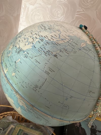 The image shows a close-up view of a vintage globe, presenting a portion of East Asia, including parts of Russia, Japan, the Korean Peninsula, and Southeast Asia. The globe appears to be an older model, with names written in Cyrillic and other languages. A beaded necklace is draped on the globe's top edge. The background reveals a patterned wall, a partial view of a framed picture, and what seems to be a woven basket holding other items. The exact location in Sayansk, Russia, cannot be determined.