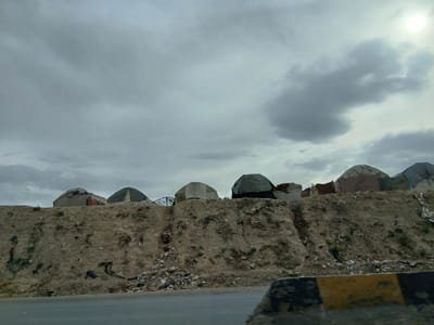 The image depicts a roadside scene in Kuchlak, Pakistan. A series of semi-permanent dwellings, possibly yurts or tents, sit atop a low, eroded hill or embankment. The hillside is largely barren with sparse vegetation and scattered debris. A significant amount of trash and refuse is accumulated at the base of the hill. The sky is overcast with heavy clouds. A concrete barrier with yellow and black markings is visible in the foreground along the road. The image is taken from a low vantage point, suggesting it was photographed from a moving vehicle. There are no people visible in the image.