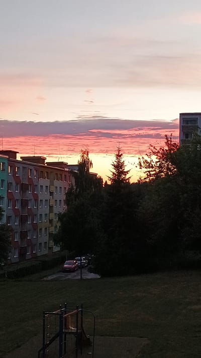A vertical, medium shot captures a view of a residential area at dusk in Sokolov, Czechia. In the foreground, a dark, grassy playground area is visible, featuring a small, blue and red climbing structure with a slide. In the mid-ground, a line of dark green trees forms a dense border. Beyond the trees, a row of apartment buildings runs horizontally across the frame. The buildings are multi-story and have a colorful, patterned facade with many windows. Several parked cars are visible in front of the buildings, including a red car. The sky above is a gradient of soft blues and pinks, with horizontal bands of clouds reflecting the sunset. The overall scene depicts a tranquil evening in a city neighborhood.
