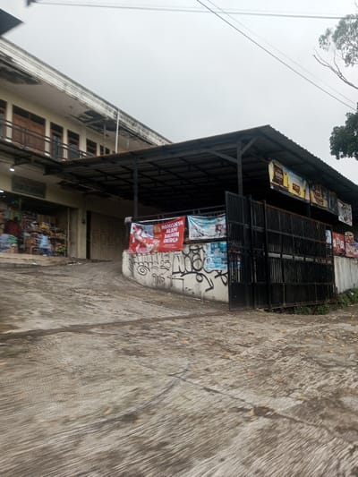 The image displays an outdoor scene in Kandapola, Sri Lanka, featuring buildings, a covered structure, and a concrete ground under an overcast sky.
To the left, a two-story building with a light-colored, possibly cream, facade stands. The upper floor features a series of identical dark wooden doors, each with a narrow balcony railing. The roofline shows some discoloration and trim. Below this, the ground floor includes a recessed storefront on the far left, appearing to be a retail shop with various items displayed and several blurred figures visible inside. Adjacent to this shop, two metal roll-up doors are closed, indicating other commercial units.
To the right of the main building, a separate covered structure extends forward. It has a dark, corrugated metal roof supported by a dark metal framework. Beneath this roof, a concrete retaining wall descends towards the left, its surface covered with black graffiti tags. Several banners are attached to this wall and the adjacent structure; a prominent red banner on the left reads "MANISNYA ALAM BIKIN ENERGI." Further to the right, a dark metal gate with vertical bars partially obscures the view beneath the covered area. Above the gate, under the roof, additional rectangular advertisements are visible. A tree with sparse foliage is partially visible against the sky on the far right.
The ground in the foreground is a textured concrete surface, showing signs of wear and faint patterns, possibly from vehicle traffic or cleaning. Scattered dark specks are visible on the concrete. The sky is uniformly overcast, and thin utility lines traverse the upper portion of the frame. No clear interactions between individuals are discernible.