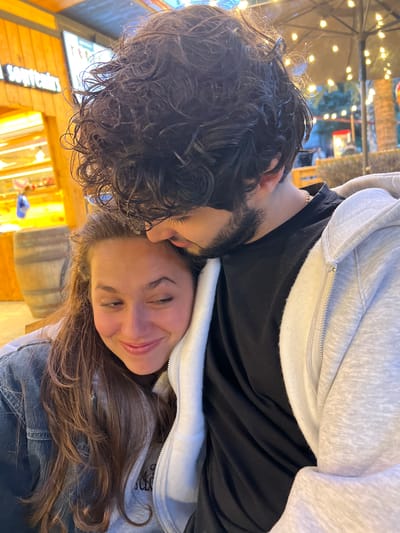 A man and woman are embracing in an outdoor setting. The woman is smiling, with brown hair and fair skin. She is wearing a denim jacket and light-colored hooded sweatshirt. The man has dark, curly hair and a beard and is wearing a black shirt and light-colored hooded sweatshirt. They appear to be close together. The background includes a barrel, wooden structures, and blurred lights. The scene appears to be taken in the city of Tsakhkadzor, Armenia.