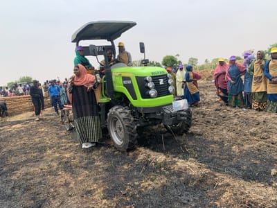 The image shows a green tractor with a black "WOFAN" logo on the side, parked on a field in Ringimawa, Nigeria, at coordinates 11.666 latitude, 8.415 longitude. The tractor is surrounded by a crowd of people. A woman wearing a blue head scarf and a black and white dress is holding a microphone and speaking. She is standing in front of the tractor. To the left of the tractor, many people stand in a group. There are some people in uniform. To the right, a group of women stand in a line. There are some trees in the background. The field is covered in grass with patches of black ash. The ground is muddy. The sky is clear and light blue.