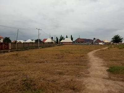 This is a wide-angle, eye-level outdoor shot of a residential area in Uke, Nigeria. The sky is overcast with grey clouds. In the foreground, a dirt path winds through dry, brown grass. The middle ground features a row of single-story houses with diverse roof designs, including gabled, hip, and flat roofs, some of which have solar panels. There is a low concrete wall bordering the houses and a rusty metal fence on the far left. Utility poles with electrical wires are visible. A few people are standing near the houses, and a white van is parked in front of one. In the background, more houses and trees are visible, along with a large, dark green coniferous tree on the right. The overall impression is one of a developing residential neighborhood.