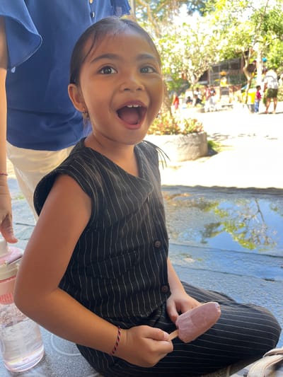 The image shows a young girl sitting down outdoors, holding a pink popsicle. She is wearing a black and white striped top and has a red, white, and black bracelet on her wrist. She is looking at the camera with her mouth open, appearing happy and excited. To her left, a person wearing a blue shirt is visible. In the background, there is a body of water that reflects trees and the sky, suggesting it is a sunny day. There are also some buildings and greenery visible in the distance. The ground appears to be a paved walkway or patio. The overall impression is of a casual outing in a tropical or subtropical location.