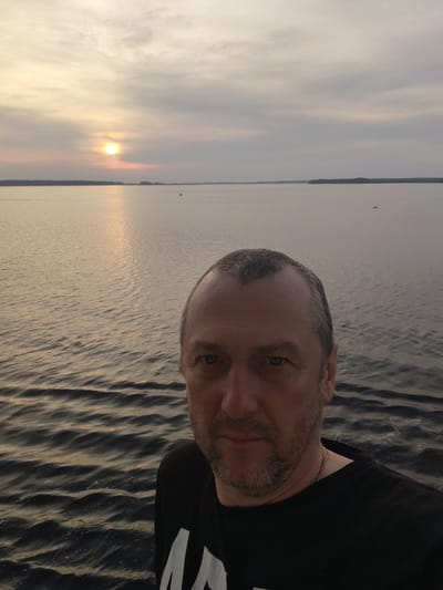 A selfie photograph taken in what appears to be Суда, Russia at sunset. The foreground contains a man wearing a black t-shirt and gold chain, centered and facing the camera. The background shows a calm body of water, presumably the Suda River or a nearby lake, reflecting the setting sun. The sky is overcast with clouds. There is land visible on the horizon.