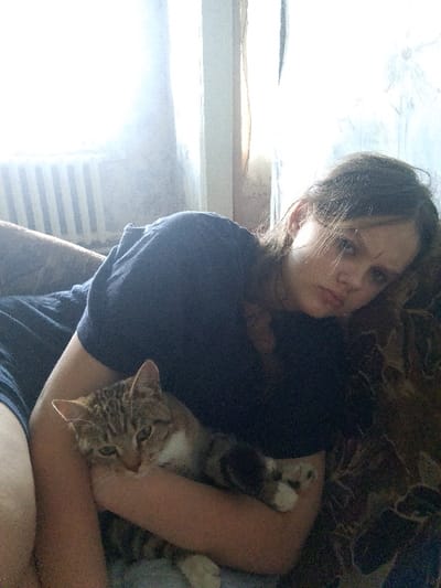 A young woman with long brown hair leans on her side, holding a tabby cat. She is wearing a dark t-shirt. The cat is nestled in her arms, looking towards the camera with its head resting on her arm. They are indoors, with a window and a radiator visible in the background. The lighting suggests it is daytime, with sunlight filtering through the window. The setting appears to be a casual domestic environment.