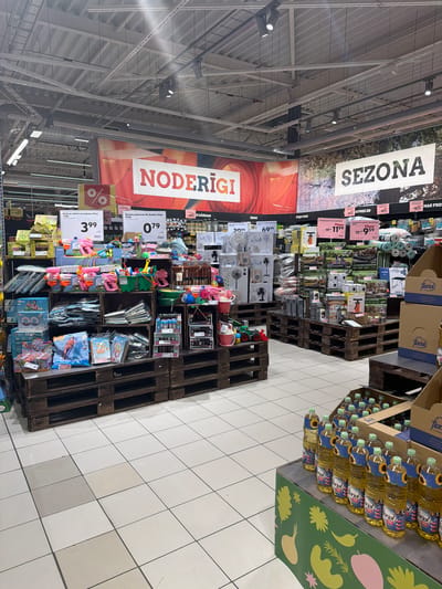 This image depicts the interior of a supermarket, specifically an aisle featuring promotional and seasonal goods, located in Krustkalni, Latvia. The scene is devoid of people.
In the foreground, to the right, a green display stand with stylized vegetable graphics holds multiple clear plastic bottles of Floriol brand cooking oil, each featuring a sunflower label. The bottles are arranged in rows. The floor consists of light-colored square tiles, some showing slight discoloration or variation in shade.
The midground is dominated by large product displays constructed from dark wooden pallets and crates. On the left side, an extensive array of children's toys and summer recreation items is presented. Visible products include pink and orange water guns, various sand play sets with buckets and shovels, inflatable pool toys (such as a "Giant Horse Ride On"), and packaged puzzles. White price tags, some displaying prices like "3.99" and "0.79", and a red circular sign with a white percentage symbol, are affixed to these displays. One sign indicates "Gross ar rokturi un audumu 24cm" and another "Burbuļu pūšamais My Bubble 55ml".
Further into the midground and to the right, additional pallet displays showcase household and outdoor items. These include boxed portable fans of various sizes and designs, packages containing what appear to be inflatable boats or air mattresses, and collapsible camping tables. Price signs here show figures such as "11.99" and "9.59," alongside red discount signs stating "-40%", with some text indicating "DĀRZAM" (for garden) and "PRECĒS" (products). Rolls of green and blue matting are also visible on these displays.
The background features large banners suspended from the ceiling. A prominent red banner on the left side displays the word "NODERĪGI" in large white capital letters. To the right, another banner, set against an image of a natural outdoor landscape with trees, reads "SEZONA" in dark capital letters. Beyond these displays, standard supermarket shelving units are faintly visible.
The ceiling is industrial in style, with exposed metallic structural elements, ductwork, numerous white fluorescent light fixtures, and black track lighting with individual spotlights directing illumination downwards onto the displays.