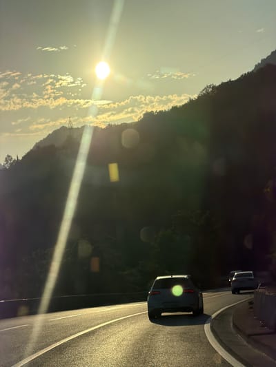 The image depicts a scenic view of a road curving through a mountainous landscape, likely near Stanišići, Montenegro, at sunset. The sun is low in the sky, casting a warm glow and creating lens flares that streak across the frame. A bright sunbeam originates from the upper left and extends diagonally downwards. The road, made of asphalt, is marked with white lines and glistens with reflected sunlight.

Several vehicles are visible on the road. In the foreground, a white hatchback car is driving away from the viewer, its rear lights illuminated. Further down the road, two other vehicles, a white sedan and possibly another car, are also moving away. The road is flanked by a steep, tree-covered mountainside on the right, and a shadowed, verdant slope on the left. A pylon for power lines is visible on the left-hand slope, silhouetted against the brighter sky. The sky itself is a gradient of pale blue and yellow, with scattered, wispy clouds catching the golden light of the setting sun. The overall atmosphere is serene and evokes the feeling of a road trip at dusk.
