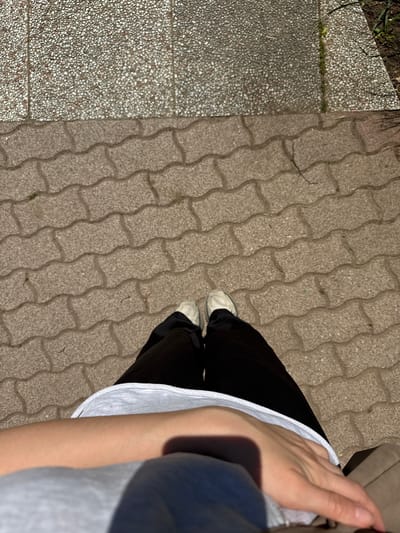 The image shows a top-down view of a person standing on a pavement in Siófok, Hungary. The pavement is made of light brown, interlocking concrete blocks. To the left is a section of textured concrete with small pebbles. The person is wearing dark pants and white shoes. A portion of a white top and an arm are visible on the right side of the image. A hand is partially obscuring the view near the upper right corner. The setting is outdoors, likely during daylight hours based on the lighting. The scene focuses on the pavement and the person's attire and immediate surroundings.
