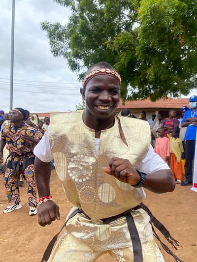In Rigachikun, Nigeria, a man is captured mid-dance, adorned in a gold vest with intricate patterns, a white t-shirt, and ornate gold pants with similar detailing. He wears a headband with shells and a wristband of red beads and shells. The man is smiling broadly, his right arm extended as if in a dynamic pose. Behind him, a crowd of people, including men, women, and children, observe. One person in the background wears a blue shirt and a face mask. The setting appears to be an outdoor gathering, with a dirt ground and a tree with green foliage overhead, suggesting a warm climate. The sky is overcast, with visible power lines and a building with a red roof in the distance.