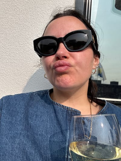 The image shows a woman in black sunglasses wearing a blue denim shirt and holding a glass of white wine. She is looking towards the camera with her lips slightly parted, giving a playful expression. She is wearing gold hoop earrings, a gold necklace, and has dark hair pulled back from her face. The image was taken on a balcony in Malmö, Sweden (latitude: 55.594, longitude: 13.01). The background is a white wall and a window with a view of a city street and a partially visible flag. The image is likely a selfie. The lighting is bright, and the image quality is high.