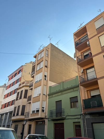 This is a low-angle, outdoor shot of buildings in Castelló De La Plana, Spain, under a clear blue sky. The sun appears to be high in the sky, suggesting it is daytime. The buildings are of various heights and colors, including beige, light green, and some with red and white striped facades. Several windows with closed blinds and some with wrought-iron balconies are visible. Antennas are mounted on the rooftops of some buildings. There is a sign on one building that reads "TAPICEROS." The perspective suggests a narrow street or alleyway between the buildings. No people or specific activities are depicted.