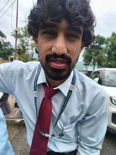 A young man with dark, curly hair and a beard, wearing a light blue collared shirt and a maroon tie, is captured in a close-up, low-angle shot. He appears to be outdoors, with a partially visible scooter to his left and a white vehicle to his right. A lanyard with the inscription "Government College Dharamshala" is visible around his neck, suggesting a connection to an educational institution. The background consists of greenery and a cloudy sky. The man is looking towards the camera with a slight smile. The lighting is natural, indicating daylight conditions.