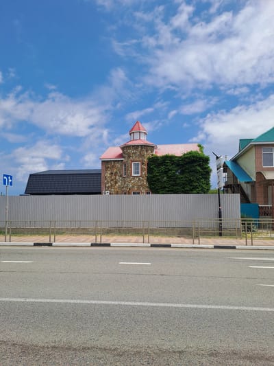 The image shows a street scene in Dzhubga, Russia. The perspective is from a low angle, looking up towards a building and the sky. The building features a stone facade with a red-tiled, conical roof and white trim. There are windows visible on the stone portion of the building. Adjacent to this structure is a building with a dark grey, tiled roof. A tall evergreen tree is visible growing up the side of the stone building.
To the right of the buildings is a paved road with white lane markings and a curb. A metal fence runs along the edge of the property next to the road. A blue traffic sign with white arrows indicating a left turn and straight ahead is mounted on a pole. The sky is blue with scattered white clouds, suggesting a daytime setting with fair weather. The overall impression is a bright, clear day. The scene appears to be a commercial or residential area bordering a road. There is no visible text in the image.